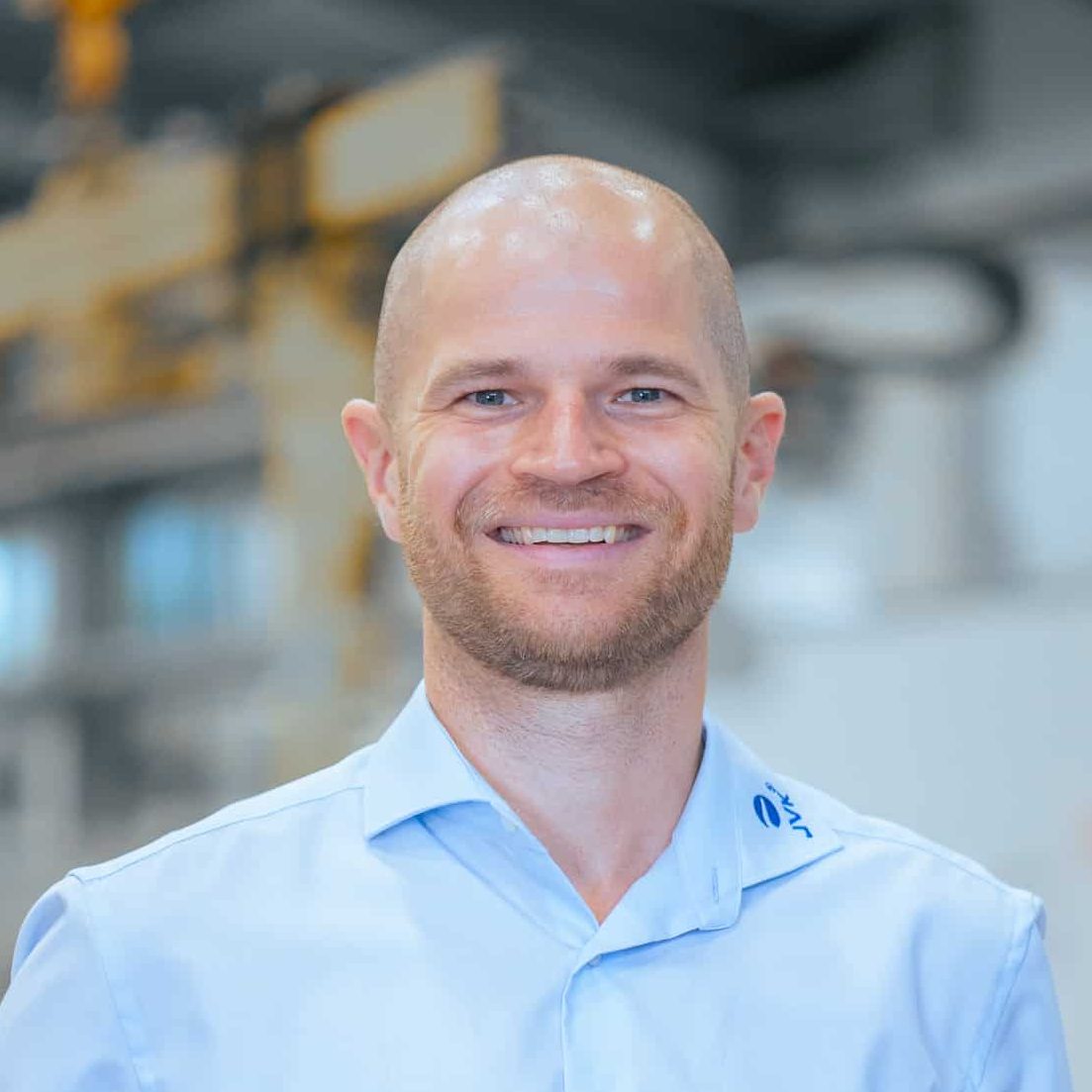Porträt eines Mitarbeiters von JVK Filtration Systems in einer modernen Produktionsumgebung. Der Hintergrund zeigt unscharf industrielle Anlagen und Fertigungstechnik.