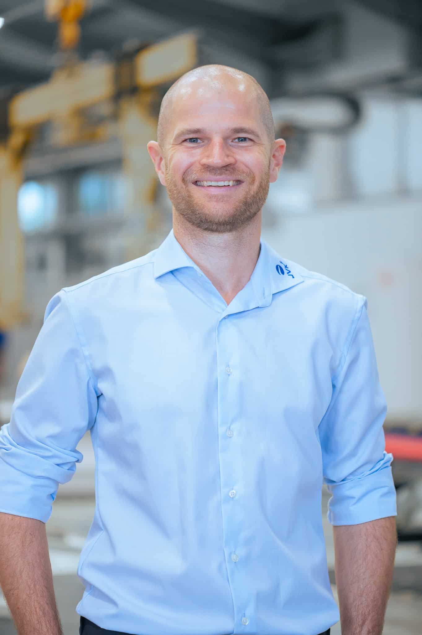 Porträt eines Mitarbeiters von JVK Filtration Systems in einer modernen Produktionsumgebung. Der Hintergrund zeigt unscharf industrielle Anlagen und Fertigungstechnik.