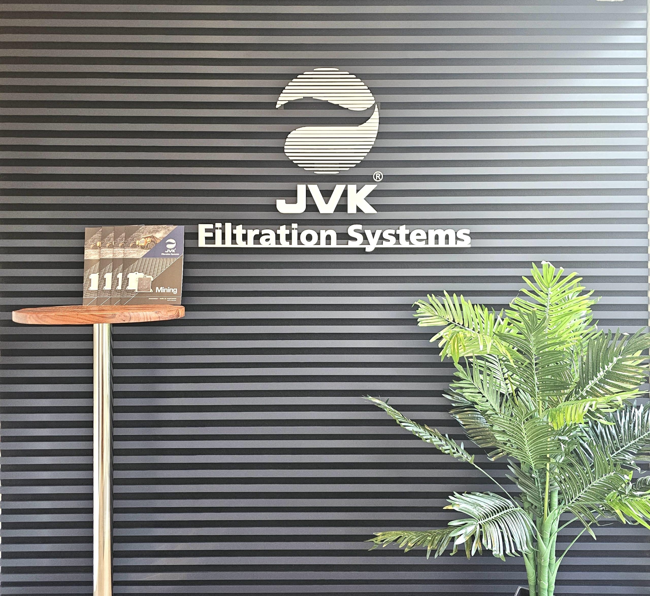 Moderne Empfangswand mit dem Logo „JVK Filtration Systems“ auf einer horizontal geriffelten, dunklen Oberfläche. Eine Pflanze und ein kleines Displayelement unterstreichen den professionellen, klar strukturierten Unternehmensauftritt.