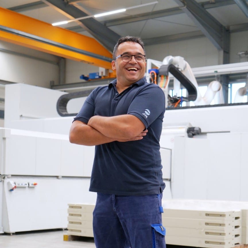 Mitarbeiter von JVK Filtration Systems steht lachend in der Produktionshalle vor modernen CNC-Maschinen. Das Bild vermittelt Teamgeist und Arbeitsfreude im industriellen Umfeld.